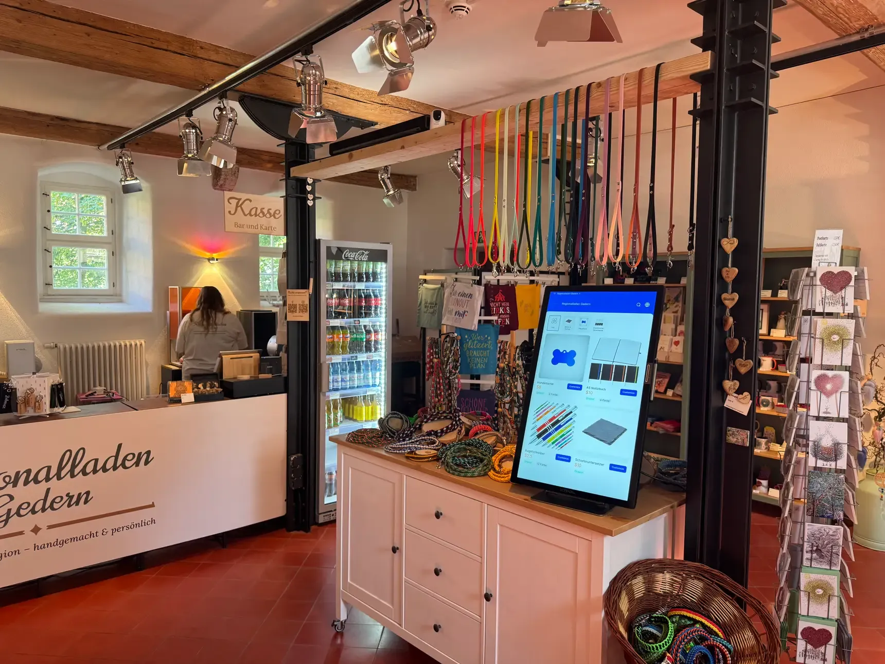 Verkaufsraum des Regionalladen Gedern: auf einer Kommode steht ein hochkant montiertes Touch-Terminal mit dem Personalisierungs-Shop, dahinter eine Wand mit farbigen Schlüsselbändern, links der Ladentresen.